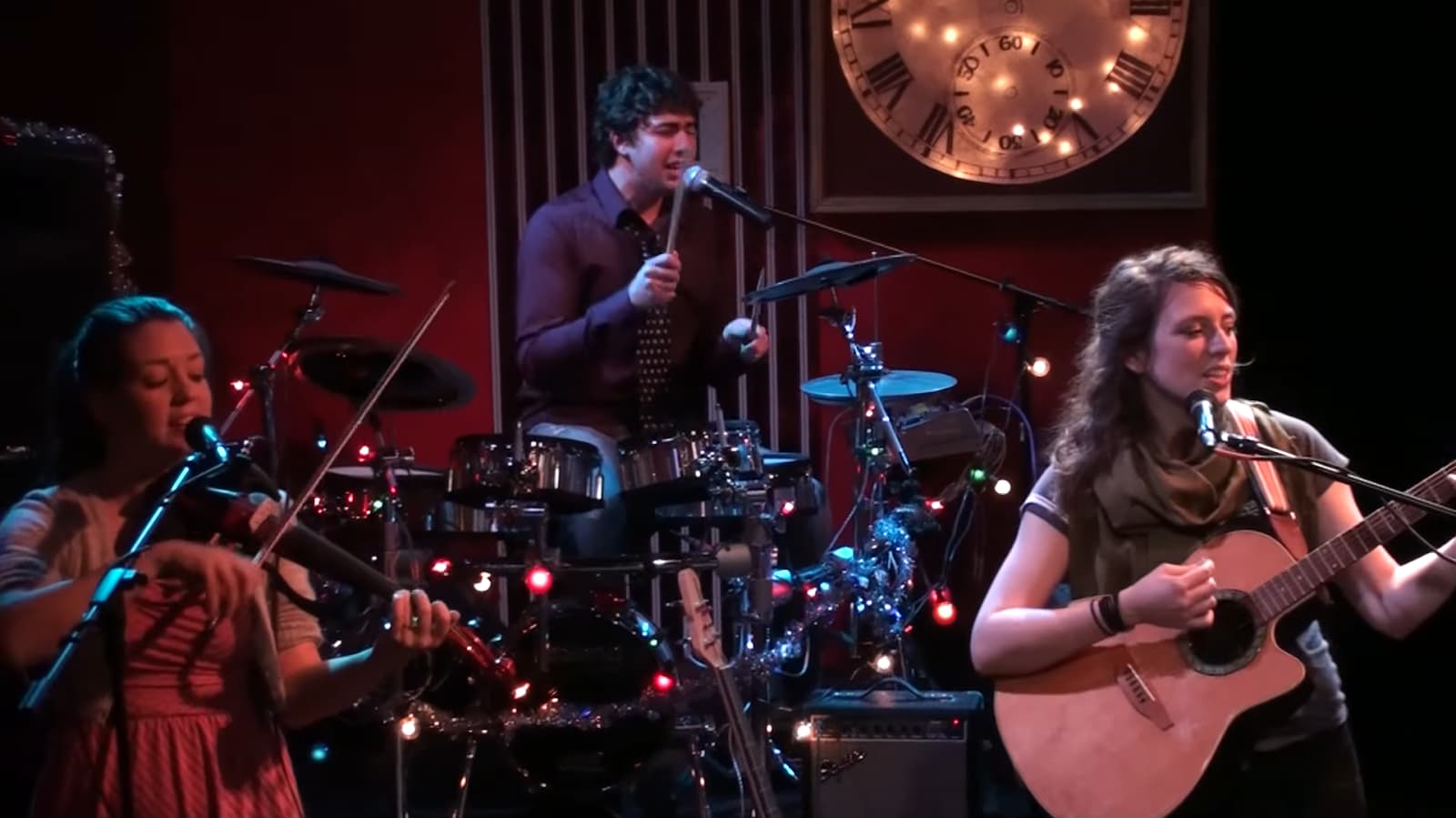 musicians perform on stage with a violin, drums, and guitar, under warm lighting