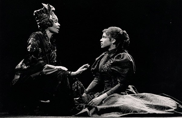 Stage scene with one actor in a fancy costume and another in period dress, having a conversation